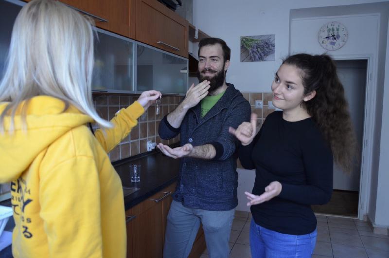 Na obrázku jsou tři postavy uvnitř prázdné kuchyně dorozumívající se znakovým jazykem