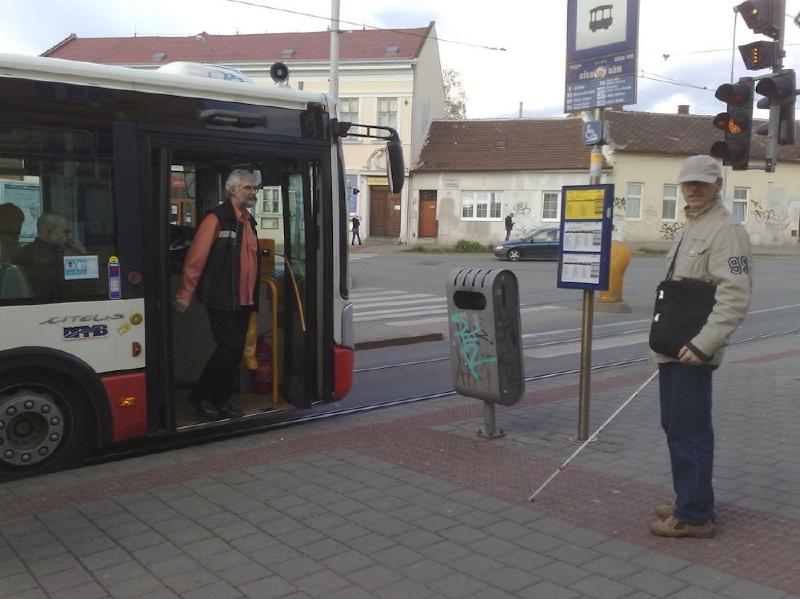 Na obrázku je nevidomý cestující u označníku zastávky. Před ním otevřené dveře trolejbusu. Nevidomý nijak nereaguje, protože neví, že trolejbus už přijel.