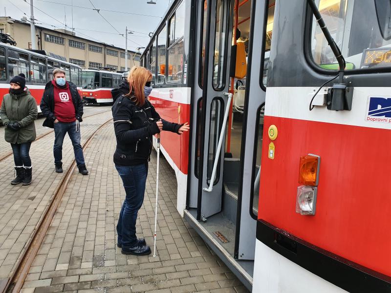 Na obrázku probíhá školení řidičů. Řidička se zaslepovacími brýlemi si zkouší nástup do tramvaje s bílou holí.